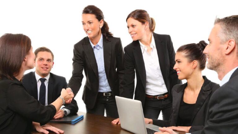 A group of six business professionals in formal attire are seated and standing around a table. Two of them are shaking hands, while others smile and look at each other, discussing vital business succession plans.