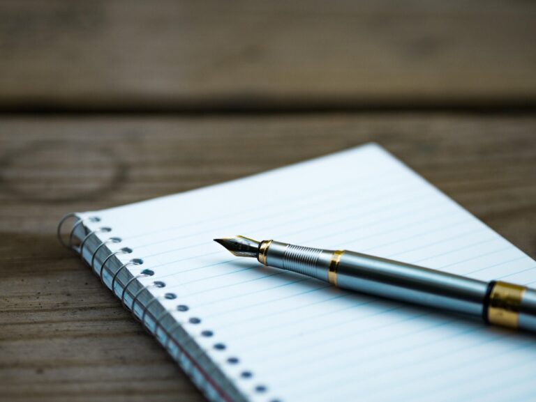 A fountain pen rests on an open spiral-bound notebook with lined paper, placed on a wooden surface, awaiting the trustee's notes.