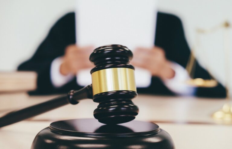 Close-up of a gavel resting on a block in the foreground, symbolizing the weight of justice, with the out-of-focus figure of a judge reading a document in the background, pondering the unintended consequences of default intestacy law.