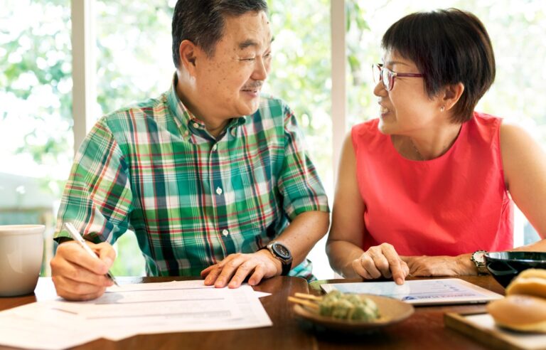 A man and woman sit at a table, reviewing documents. The woman points at a tablet, and the man holds a pen. Amidst food items and a coffee cup, they seem focused on tracing beneficiaries.