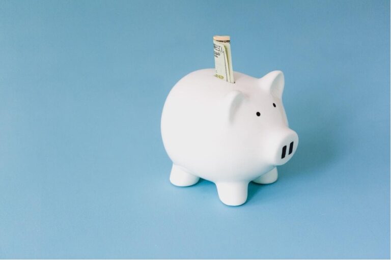 A white piggy bank with a rolled-up dollar bill inserted into the slot, set against a blue background symbolizes the importance of setting up a trust in Singapore.