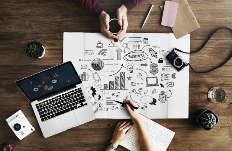 Two people collaborate over a business strategy plan on a table with a laptop, cameras, coffee cup, and notebooks, discussing essential details including CPF nomination strategies.