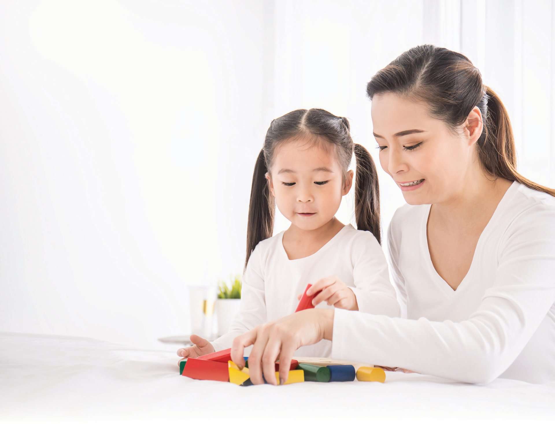 A woman and a young girl with pigtails are sitting together, playing with colorful block toys on a white surface, building not just structures but a legacy of trust and shared memories.