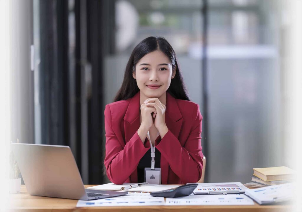 A woman in a red blazer sits at a desk with a laptop, documents, and books, looking directly at the camera with hands resting under her chin.