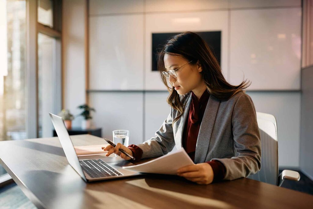 A woman in a grey blazer and glasses sits at a desk, working on a laptop and holding a document in one hand, with a glass of water nearby.