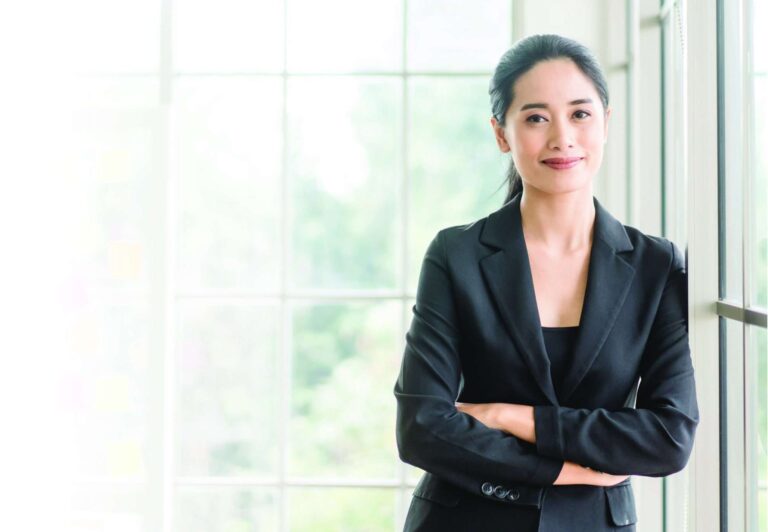 A woman in a black blazer stands by a window with her arms crossed, smiling softly.