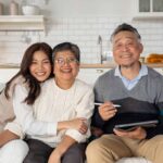 Three people sit on a couch in a kitchen, smiling at the camera. The older man holds a stylus and tablet—perhaps planning their Family Trust—while the younger woman hugs the older woman in the center, showing how they protect your wealth and legacy together.