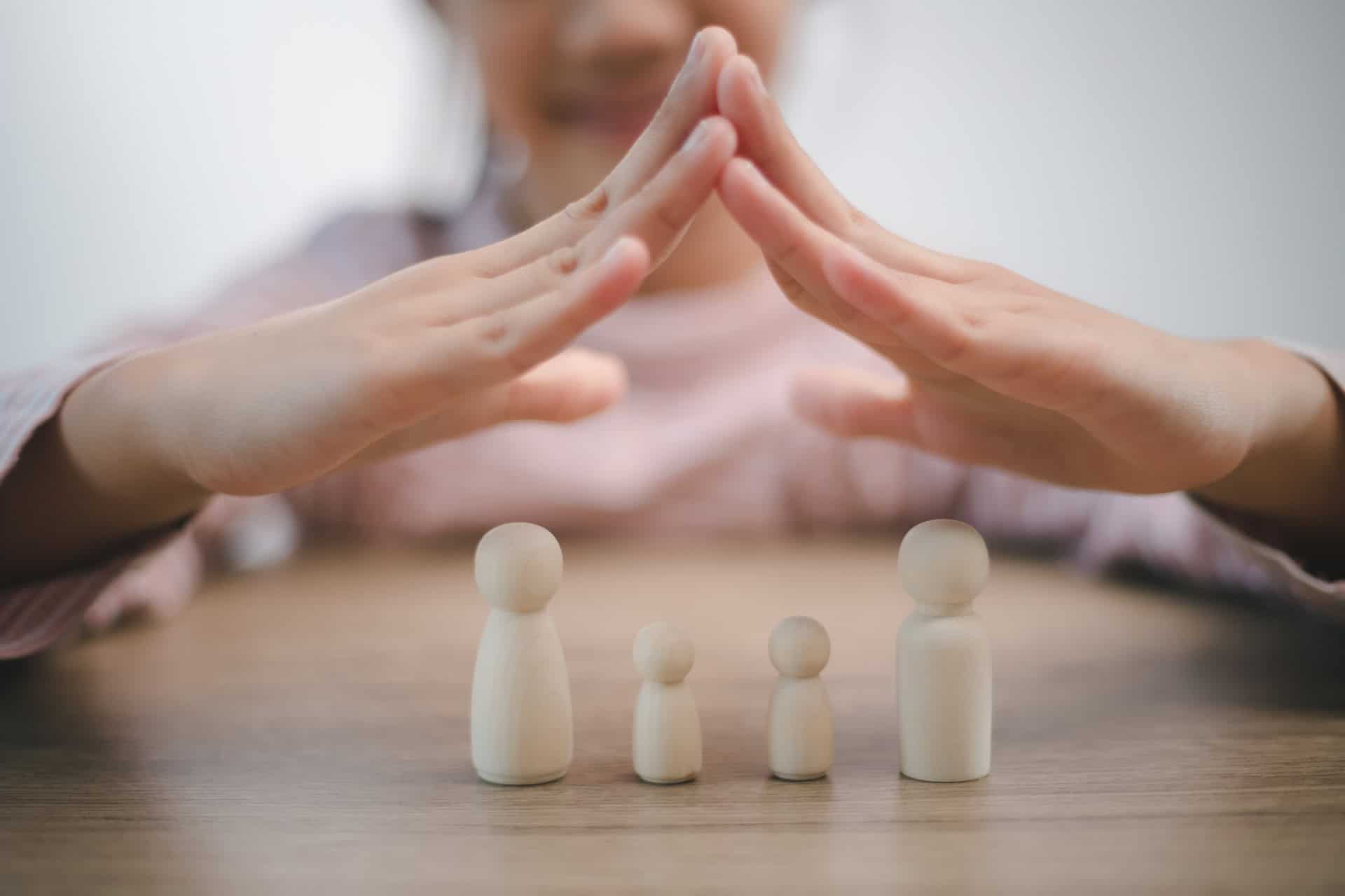 A person forms a roof shape with their hands over four wooden figurines resembling a family on a table, symbolizing how a Family Trust can help protect your wealth and legacy for future generations.