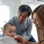 A man and a woman smile and interact with a baby who is lying on a bed in a bright room.