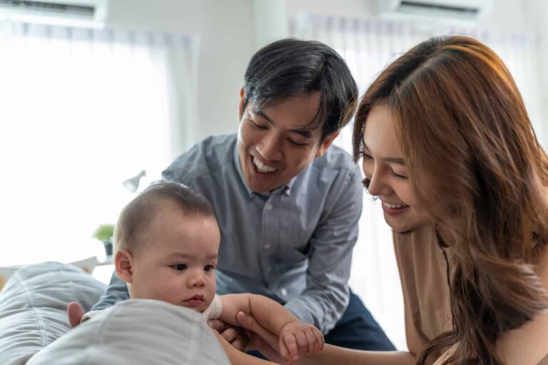 A man and a woman smile and interact with a baby who is lying on a bed in a bright room.