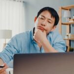A man in a blue shirt sits at a desk, looking thoughtfully at a laptop screen, holding a pen in one hand with his chin resting on the other.