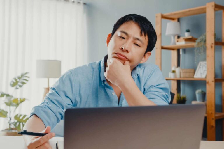 A man in a blue shirt sits at a desk, looking thoughtfully at a laptop screen, holding a pen in one hand with his chin resting on the other.