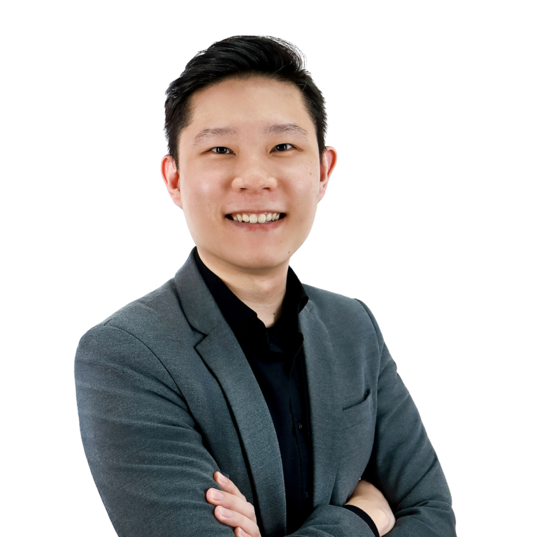 A person with short dark hair in a grey suit jacket and black shirt stands with arms crossed, smiling, against a white background.