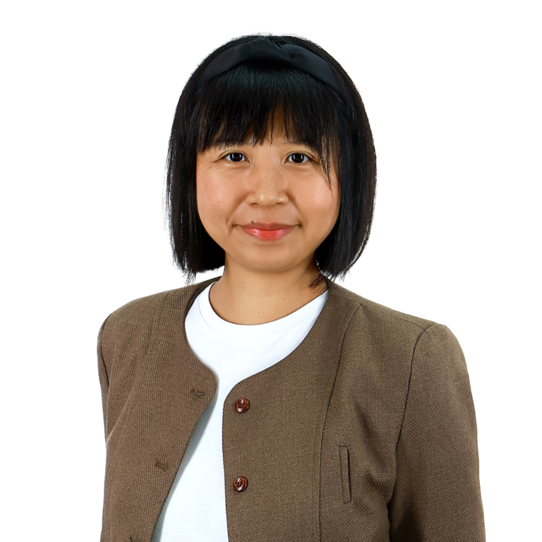 A woman with straight black hair, wearing a brown jacket over a white top and a black hairband, stands against a plain white background.