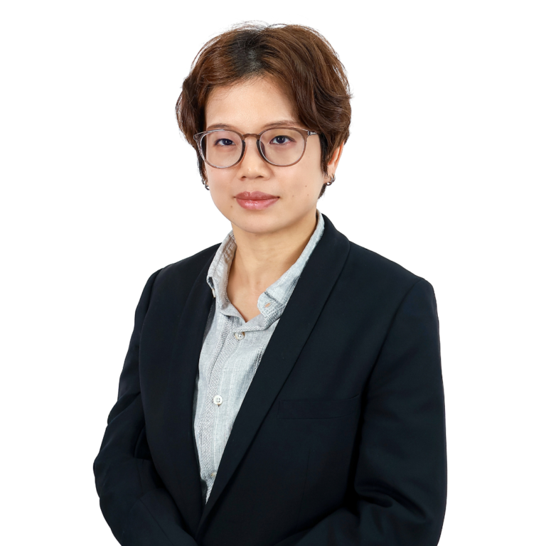 A person with short brown hair and glasses is wearing a dark blazer over a light grey shirt, standing against a plain white background.