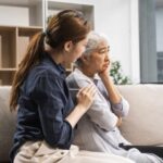 A younger woman sits beside an older woman on a sofa, offering comfort as the older woman appears upset and thoughtful, perhaps discussing sensitive topics like Trusts or SNTC.