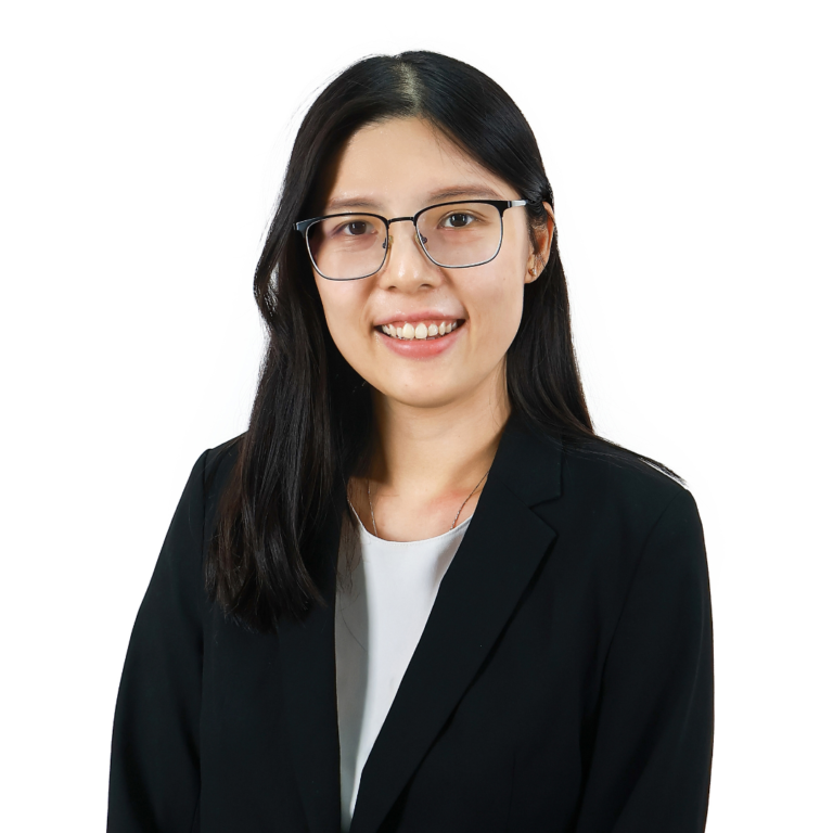 A woman with long dark hair and glasses, wearing a black blazer and white top, stands against a plain white background, smiling at the camera.