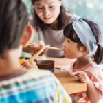 A woman, possibly a legal guardian, helps a young girl eat with chopsticks at a table, whilst another child sits nearby.